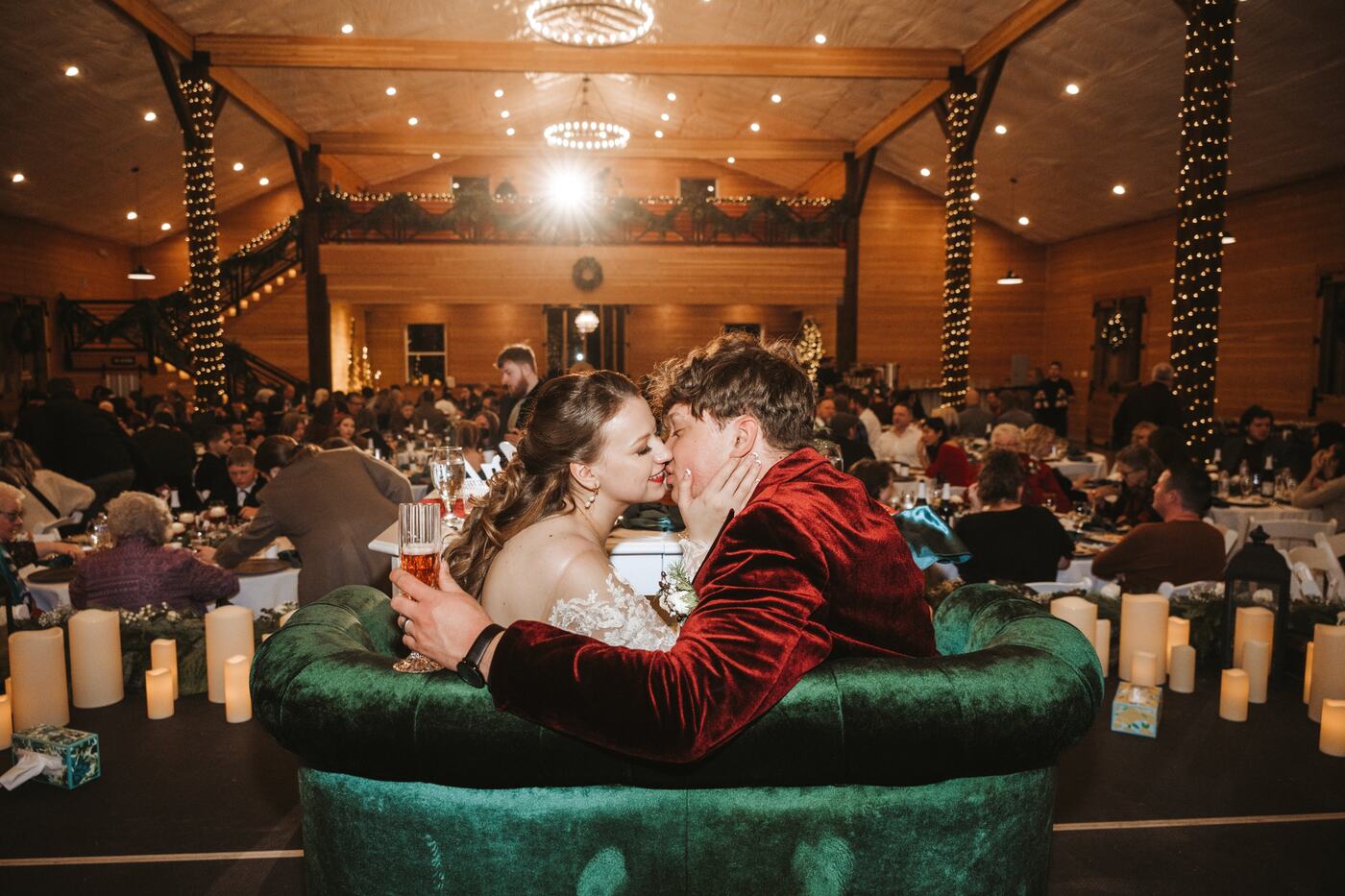 Couple kissing on a green velvet sofa at the front of the barn with the reception in full swing behind them