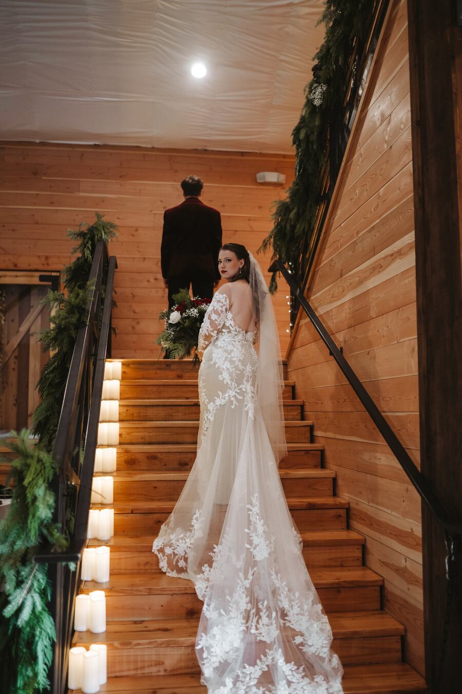 Bride looking back over her shoulder on the candlelit staircase, her lace train spilling down the steps