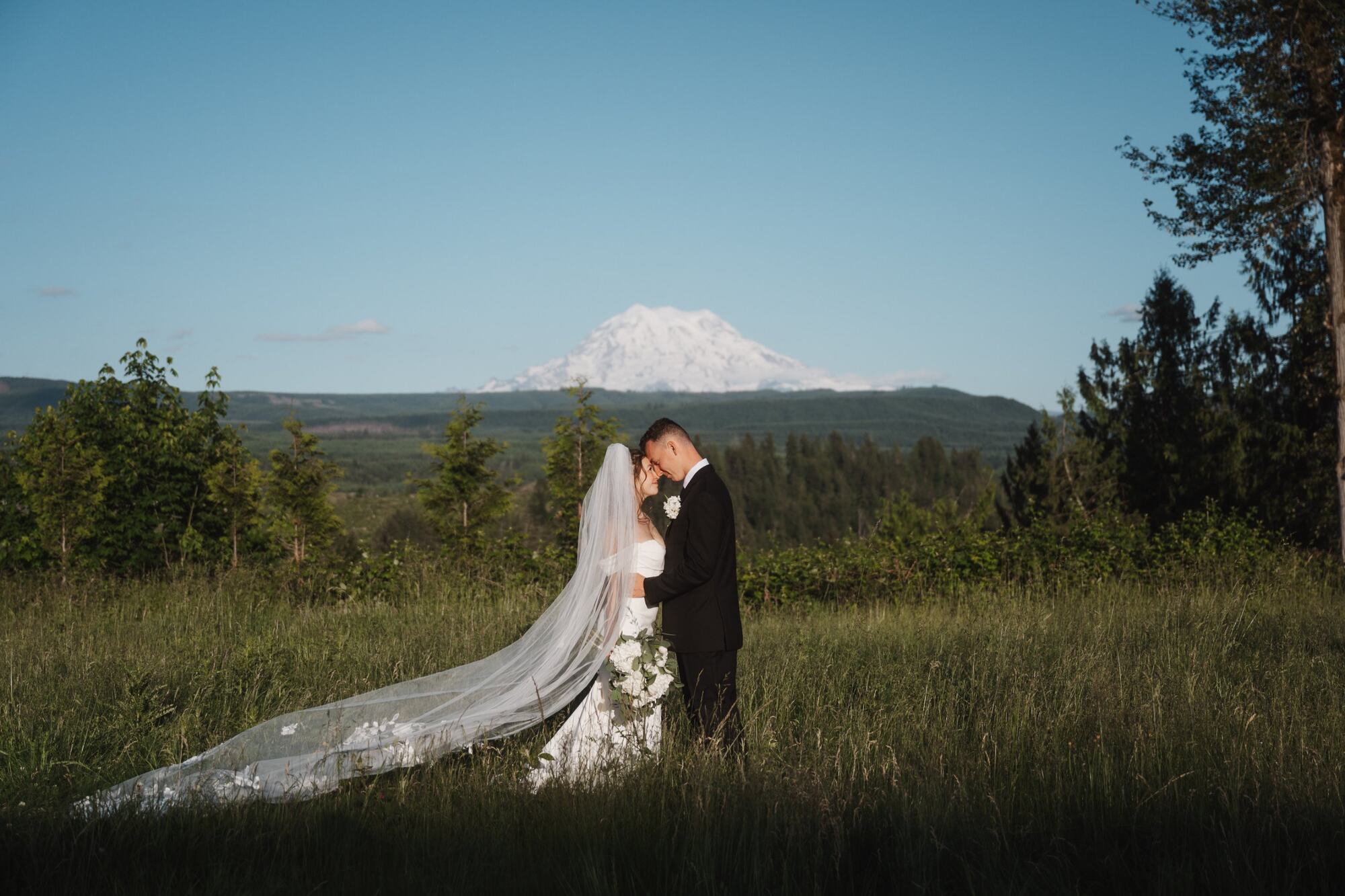A bride's veil lifting in the wind as the couple stands in the meadow with Mount Rainier behind them