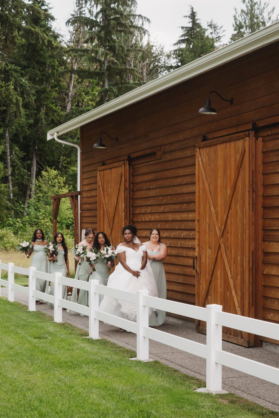 Bride and bridesmaids in sage green walking the path behind the barn along the white fence