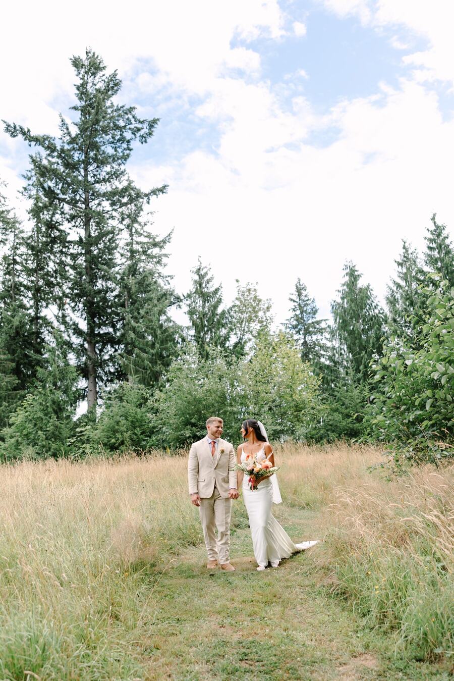 Couple walking hand in hand down a mowed path through golden grass beneath tall firs