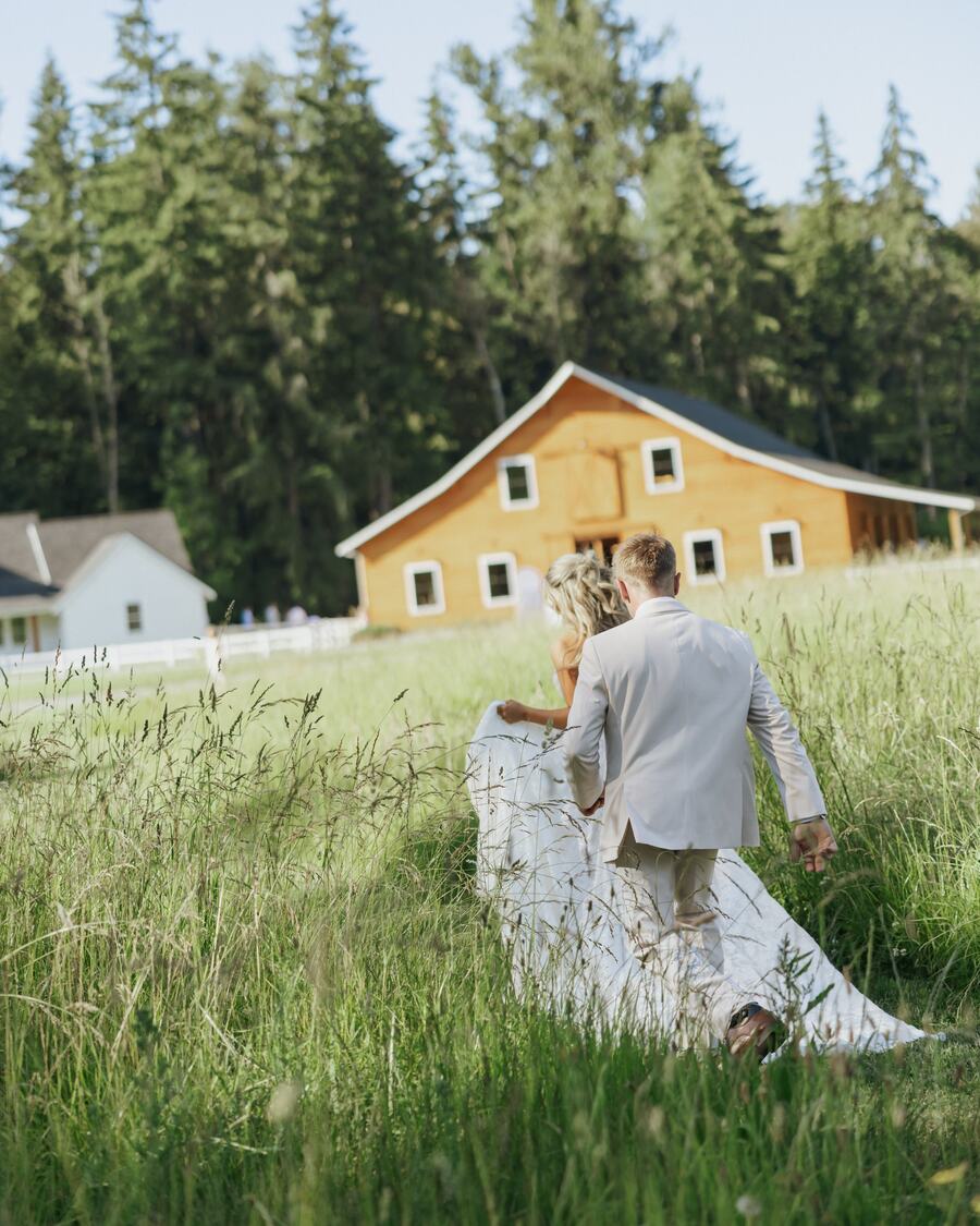 Couple wading through tall meadow grass toward the barn and bridal cottage